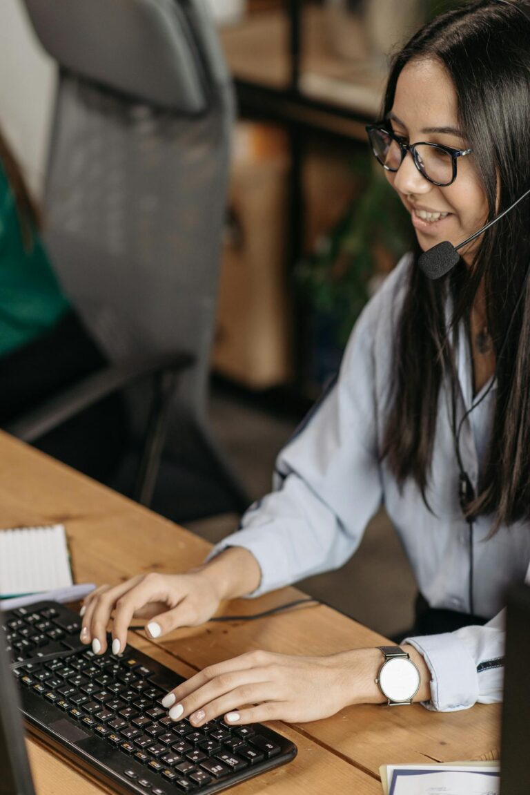 Cheerful woman in a call center typing on keyboard, wearing headset and eyeglasses.