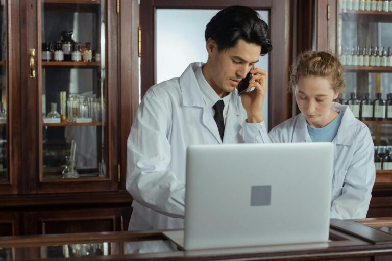 Two pharmacists working together in a pharmacy, using a laptop and phone for efficient operations.